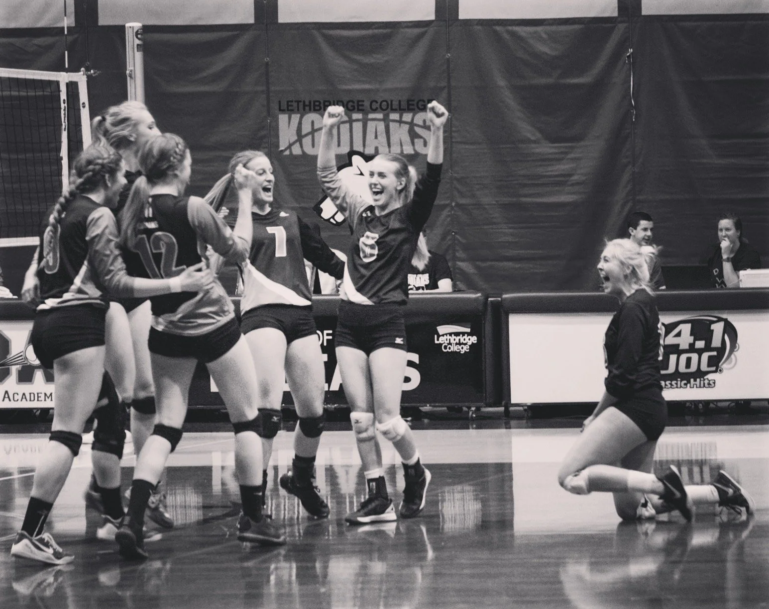 A women's volleyball team celebrating on the court, with one player kneeling and others standing, while a teammate stands with arms raised in victory, during a match at Lethbridge College.
