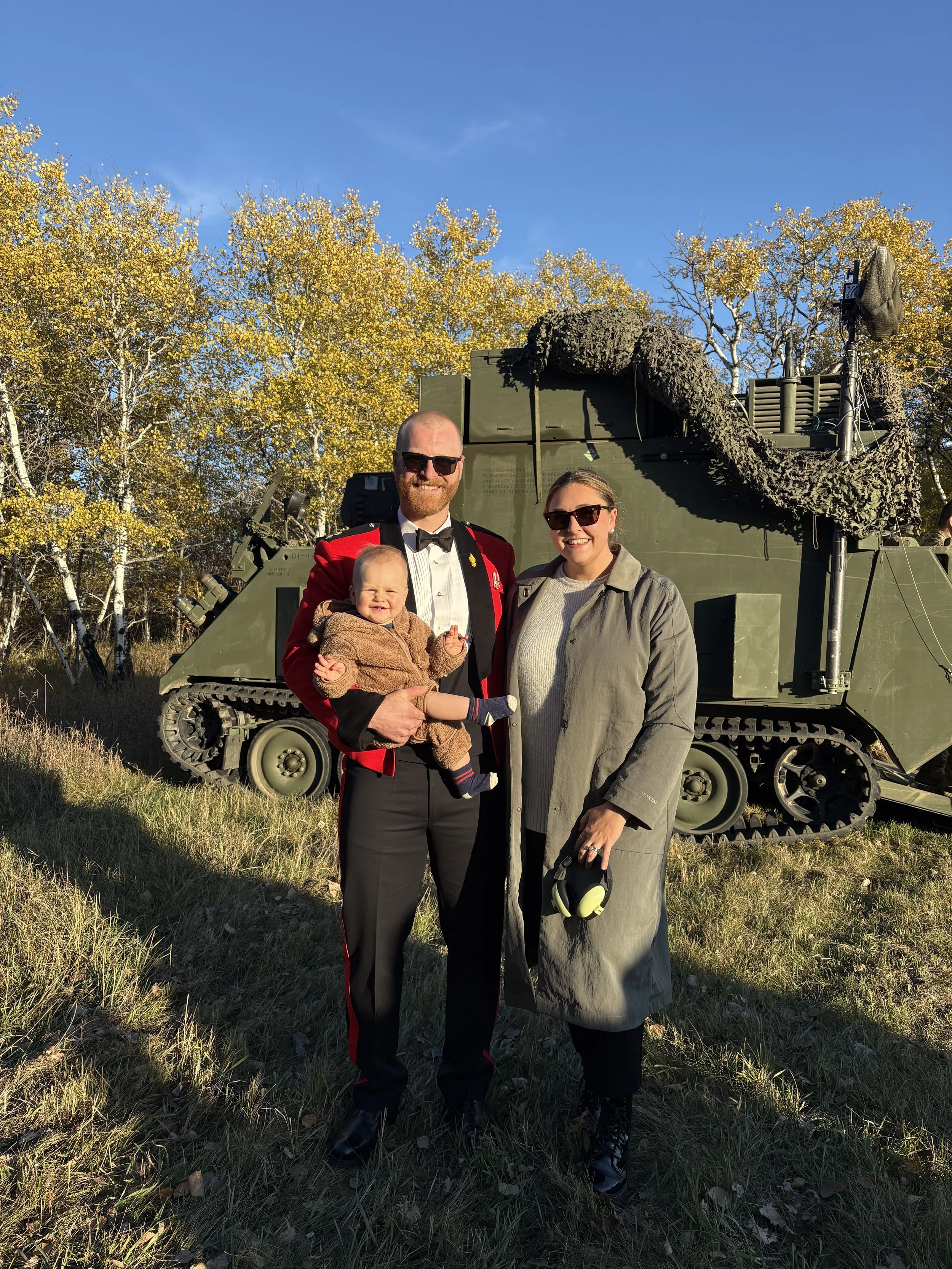 A man in a red military dress uniform, a woman in a gray jacket holding headphones, and a baby dressed in brown, standing in front of a military vehicle with a camouflage netting, outdoor setting with trees and clear blue sky.