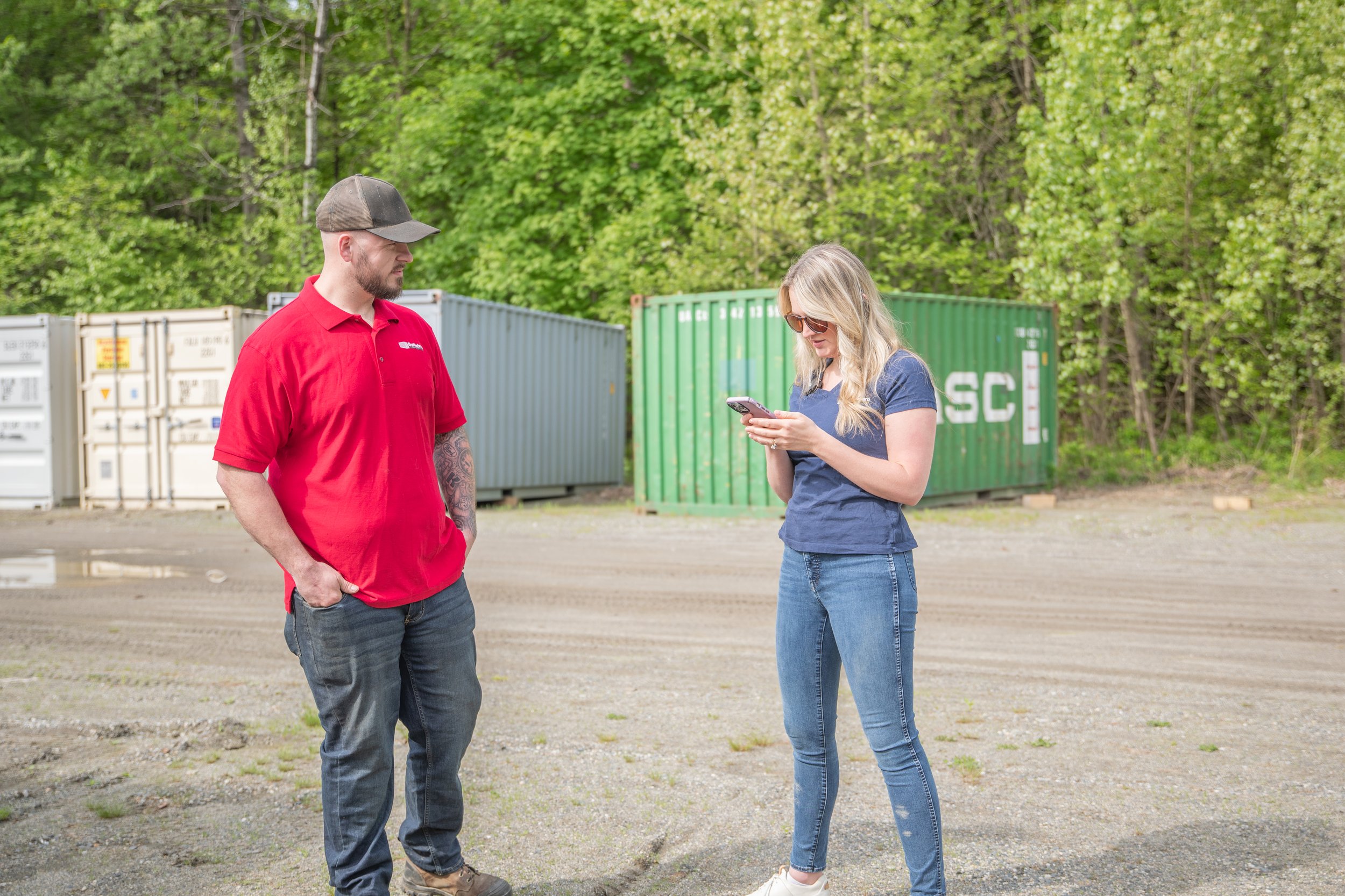 Two people, a man wearing a red polo shirt and a woman in a blue shirt and sunglasses, standing outside near containers and green trees; they are looking at a phone.
