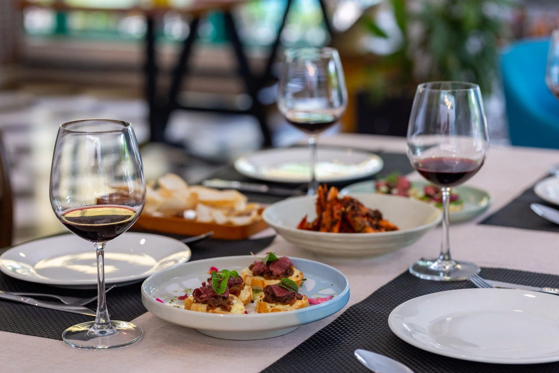 A dining table set with wine glasses, plates of appetizers including crostini with toppings, and bowls of food. The background shows a blurred indoor setting with natural light.