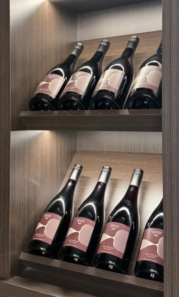 Six bottles of red wine on a wooden wine shelf, arranged in two rows with three bottles on each shelf.