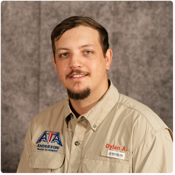 A young man with short brown hair, a goatee, and a mustache wearing a beige shirt with patches that read "ATA Trade Academy" and a name tag that says "Dylan A."; posing against a gray textured background.