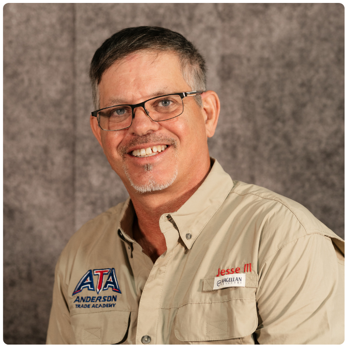 A man with short dark hair and glasses smiling at the camera, wearing a beige shirt with patches and a name tag that reads Jesse M, standing in front of a gray textured background.