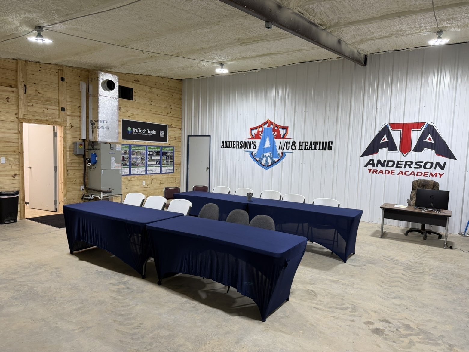 Interior of a classroom or training room with tables covered in dark blue cloth, chairs, wall logos for Anderson's A/C & Heating and Anderson Trade Academy, a desk with a computer, and wall posters.