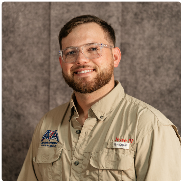 Portrait of a man with glasses and a beard wearing a beige shirt with patches, standing against a gray textured background.