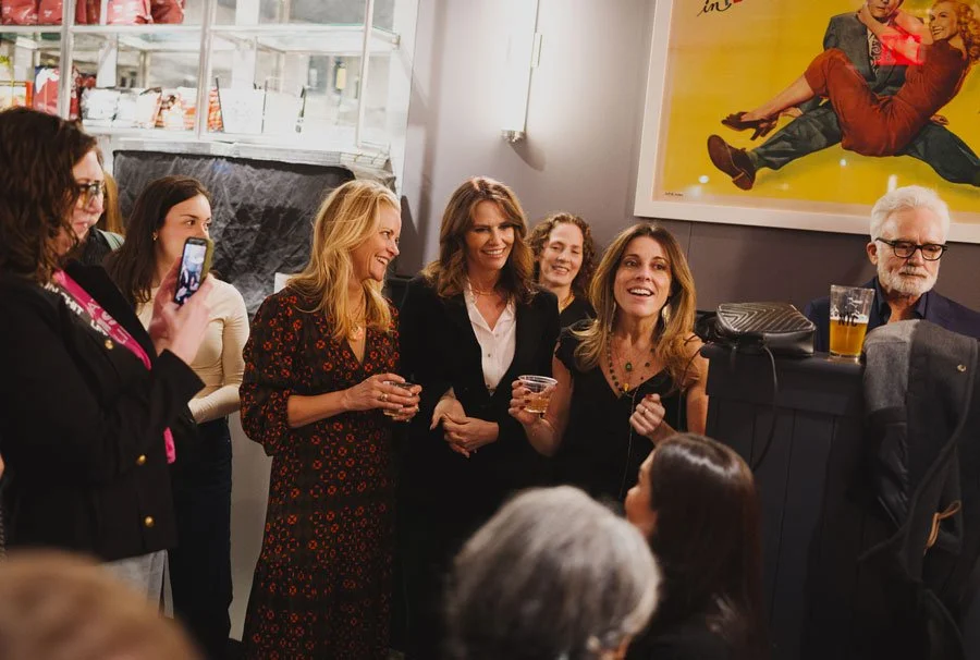 Group of women and a man gathered at a party, smiling and holding drinks, with one woman taking a photo, in a room with a poster on the wall.
