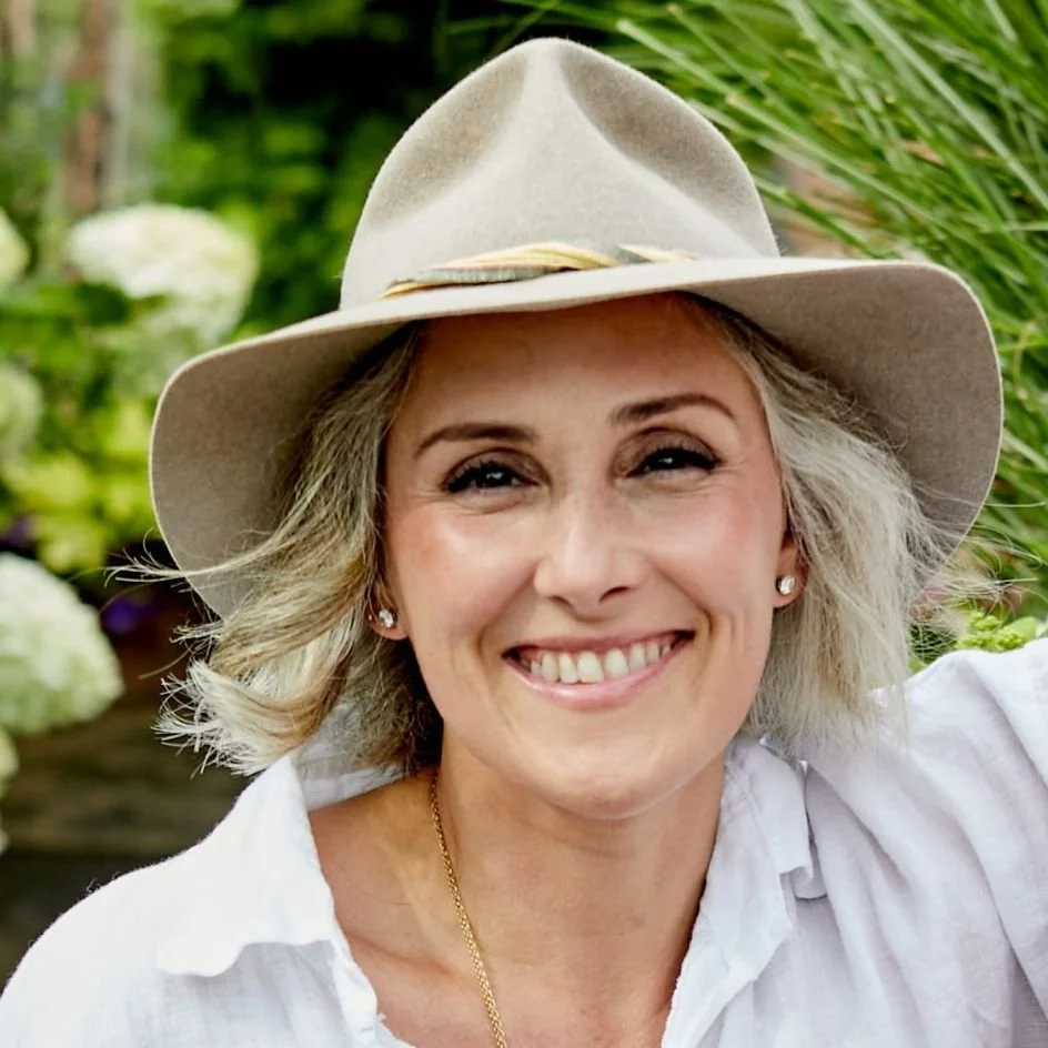 Smiling woman with short blonde hair wearing a wide-brimmed hat and white shirt, outdoors surrounded by green plants and flowers.