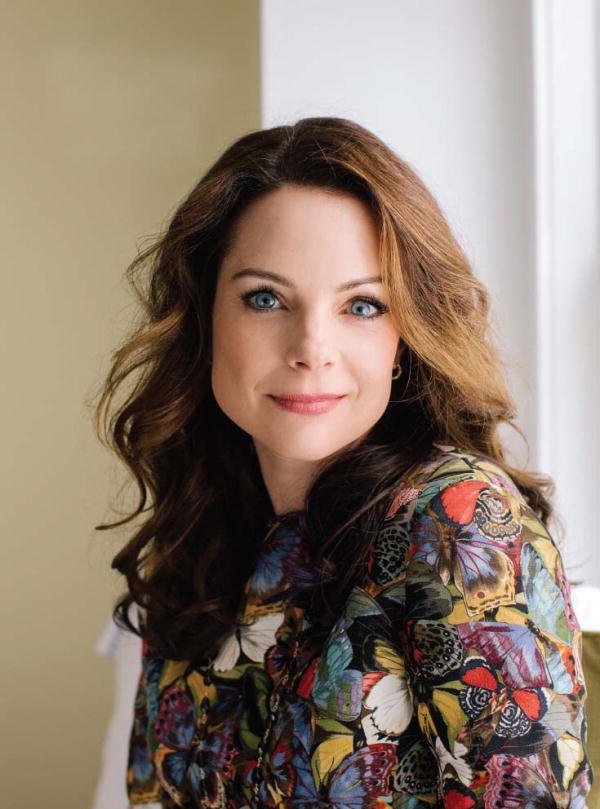 A woman with wavy brown hair and blue eyes, wearing a colorful floral blouse, smiling while looking at the camera near a window.
