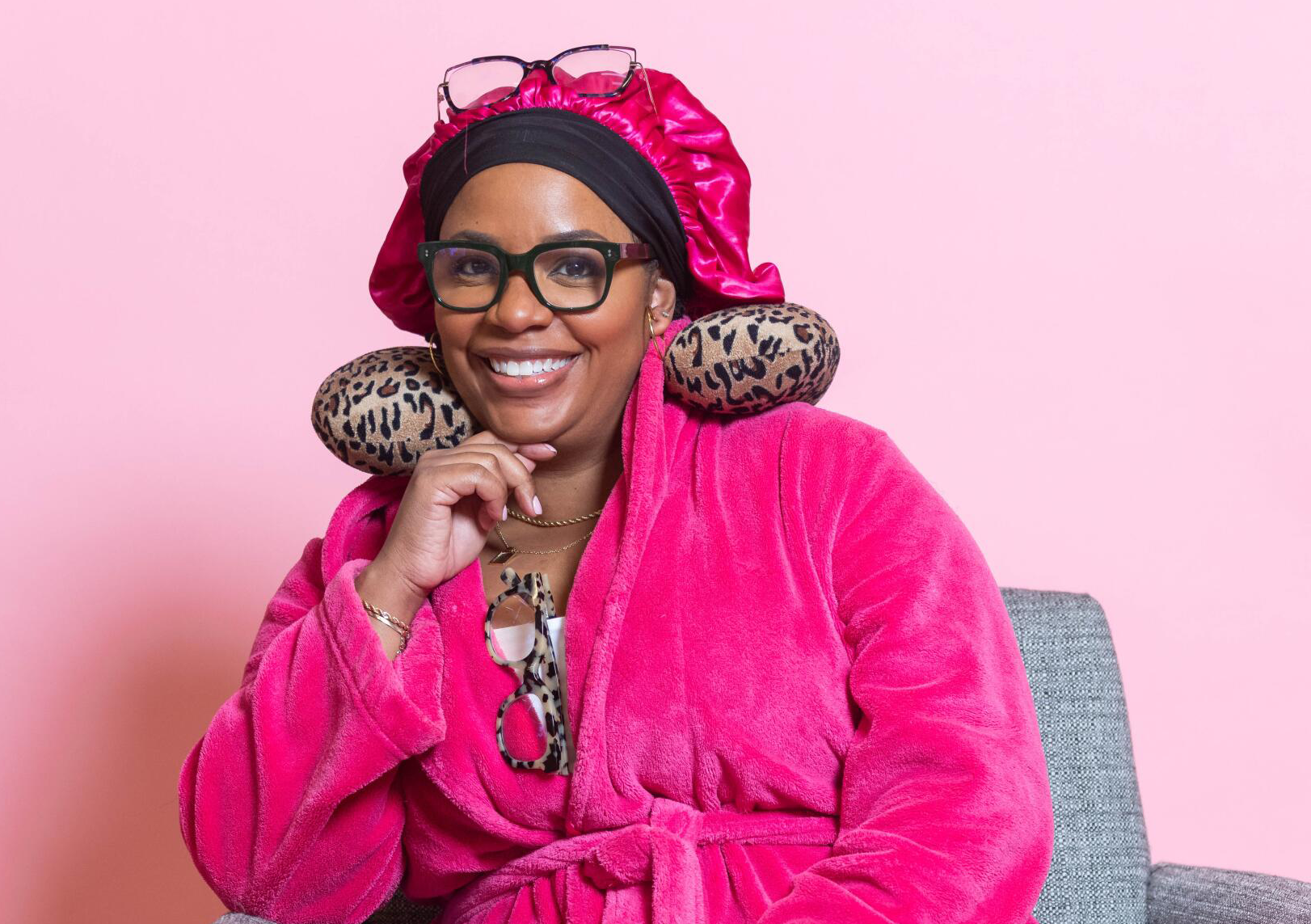 Woman wearing a pink robe, leopard print neck pillow, black headscarf, and glasses, smiling against a pink background.