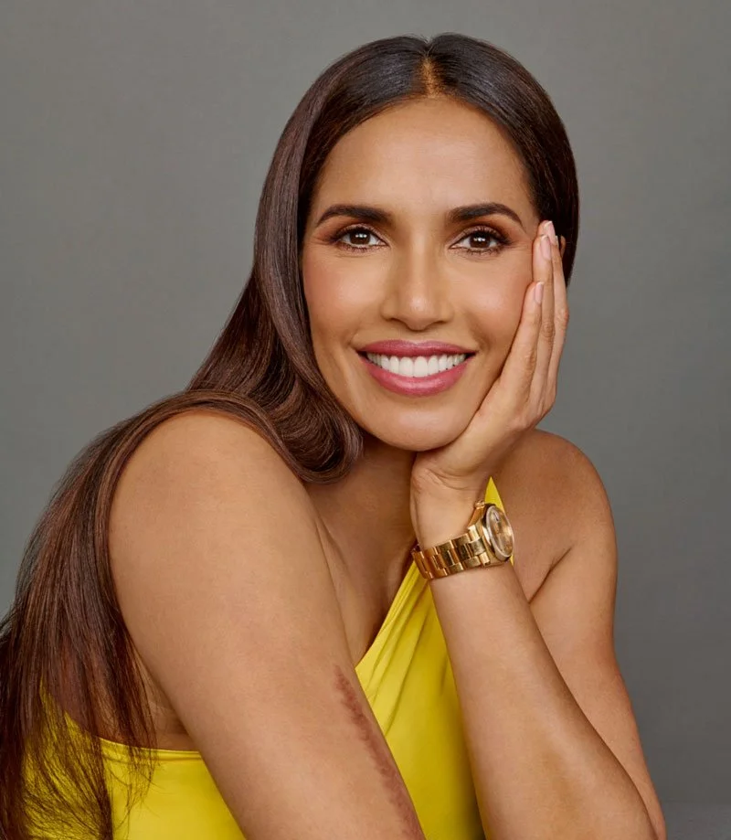 A smiling woman with long brown hair wearing a yellow sleeveless top and a gold watch, resting her hand on her face, with a visible scar on her arm, against a gray background.