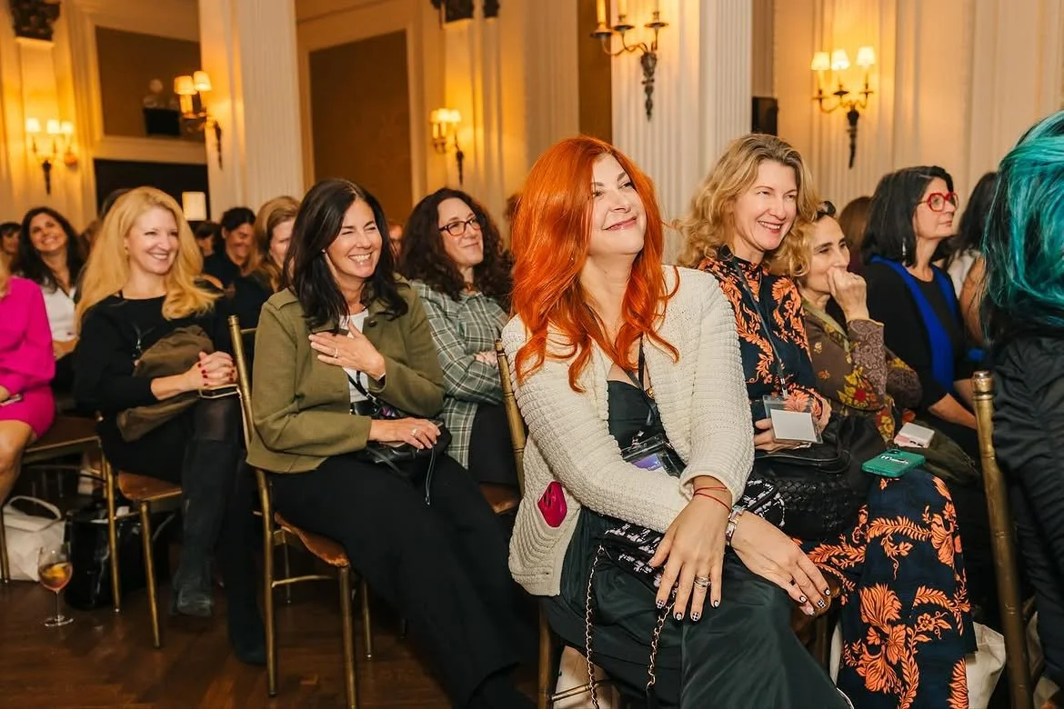 Group of women sitting in an audience, smiling and enjoying a presentation or event in a formal setting with elegant lighting.