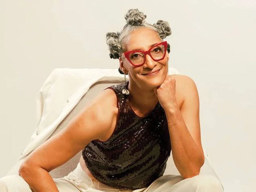 A woman with gray hair styled in bantu knots, wearing red glasses and a sleeveless black sequined top, sitting on a white chair and smiling with her chin resting on her hand.