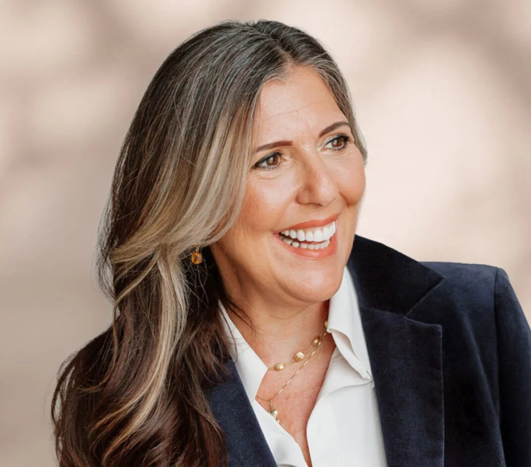 A smiling woman with long, wavy brown and gray hair, wearing a dark blazer, white shirt, jewelry, and earrings.