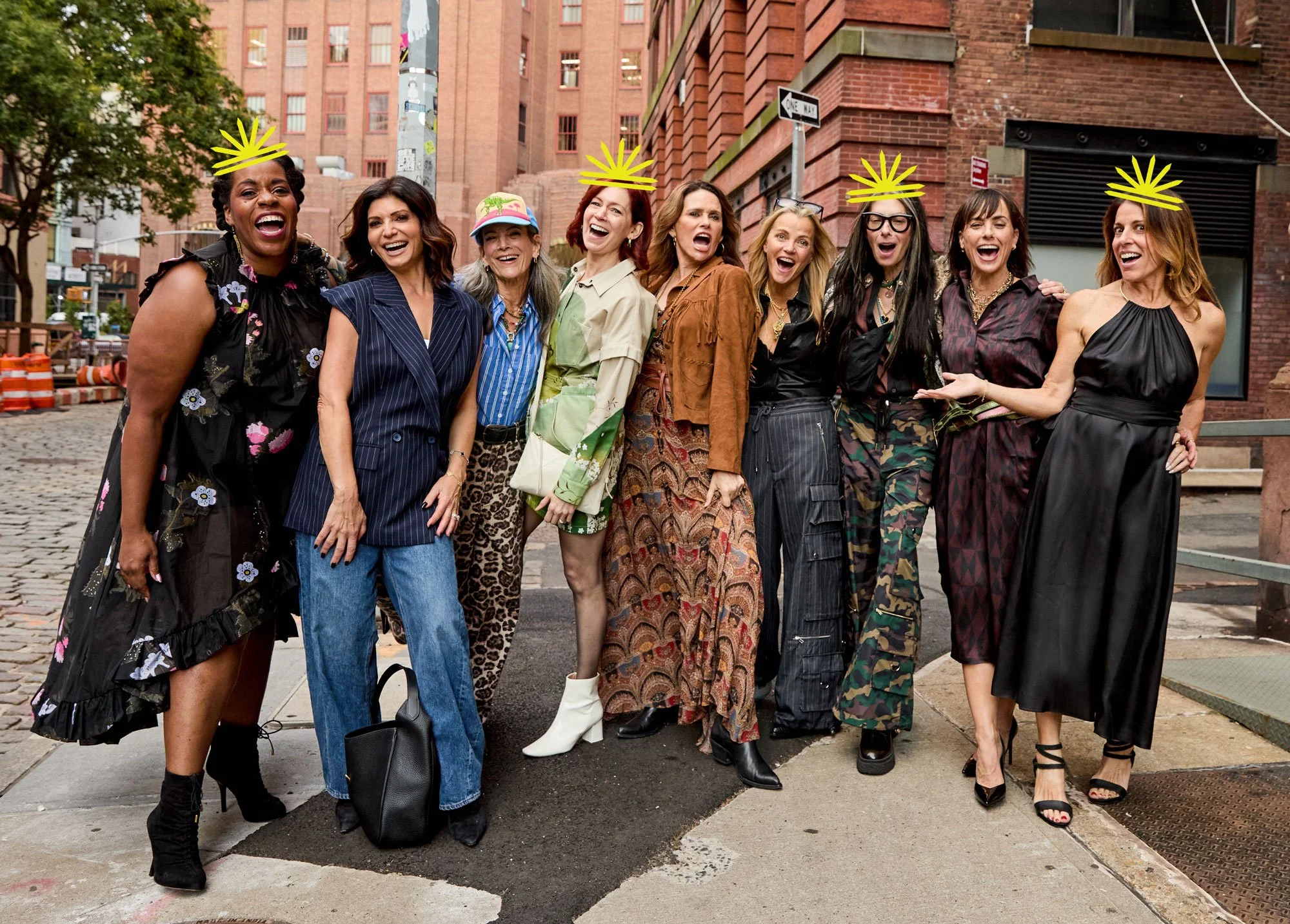 A group of ten women standing close together on a city sidewalk, smiling and laughing, with some wearing crowns. The women are dressed in a variety of fashionable outfits, including dresses, pants, and jackets. The background features brick buildings, a street corner, and street signs.