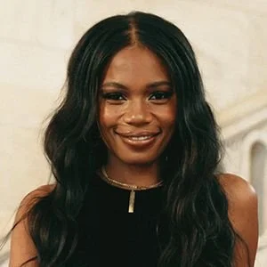 A woman with long wavy black hair smiling at the camera, wearing a black sleeveless top and a gold necklace.