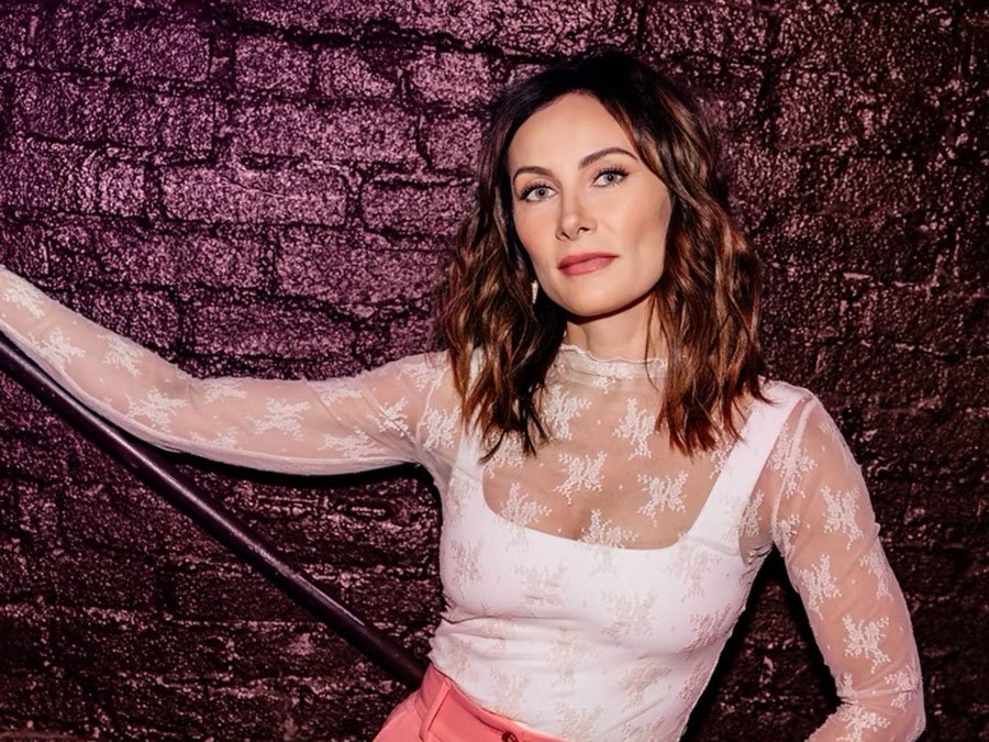 A woman with wavy brown hair in a pink and white lace top standing against a textured dark pink wall.