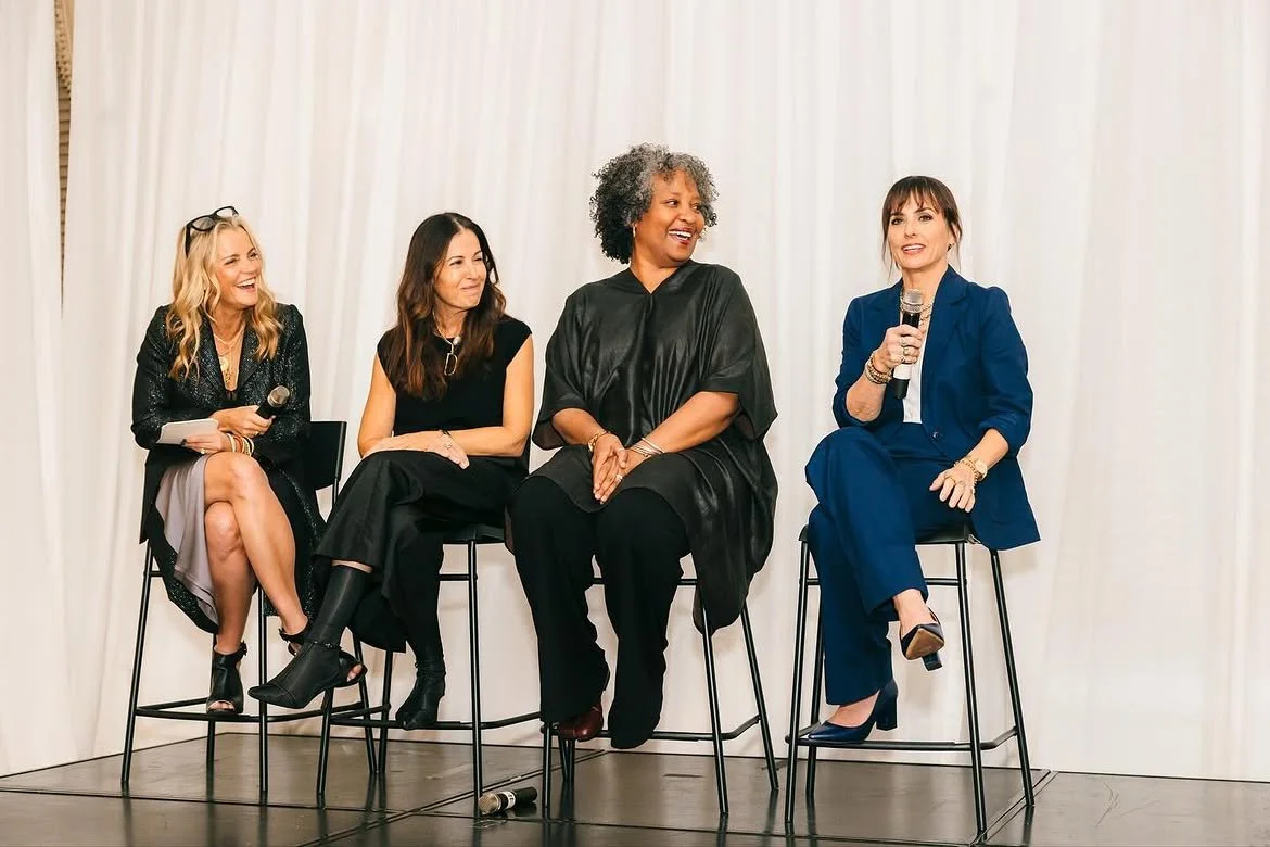 Four women sitting on high chairs on a stage, participating in a panel discussion with microphones, against a white curtain backdrop.