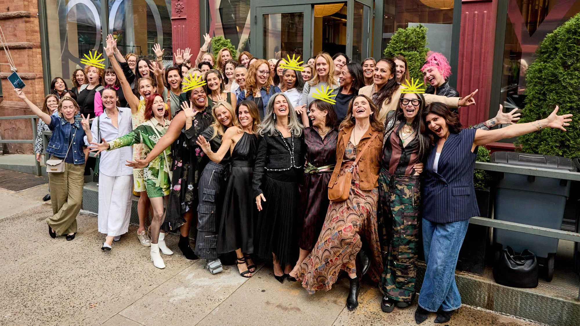 A diverse group of women celebrating outside a building with a glass entrance, some wearing yellow paper crowns, and smiling or laughing with arms raised in joy.
