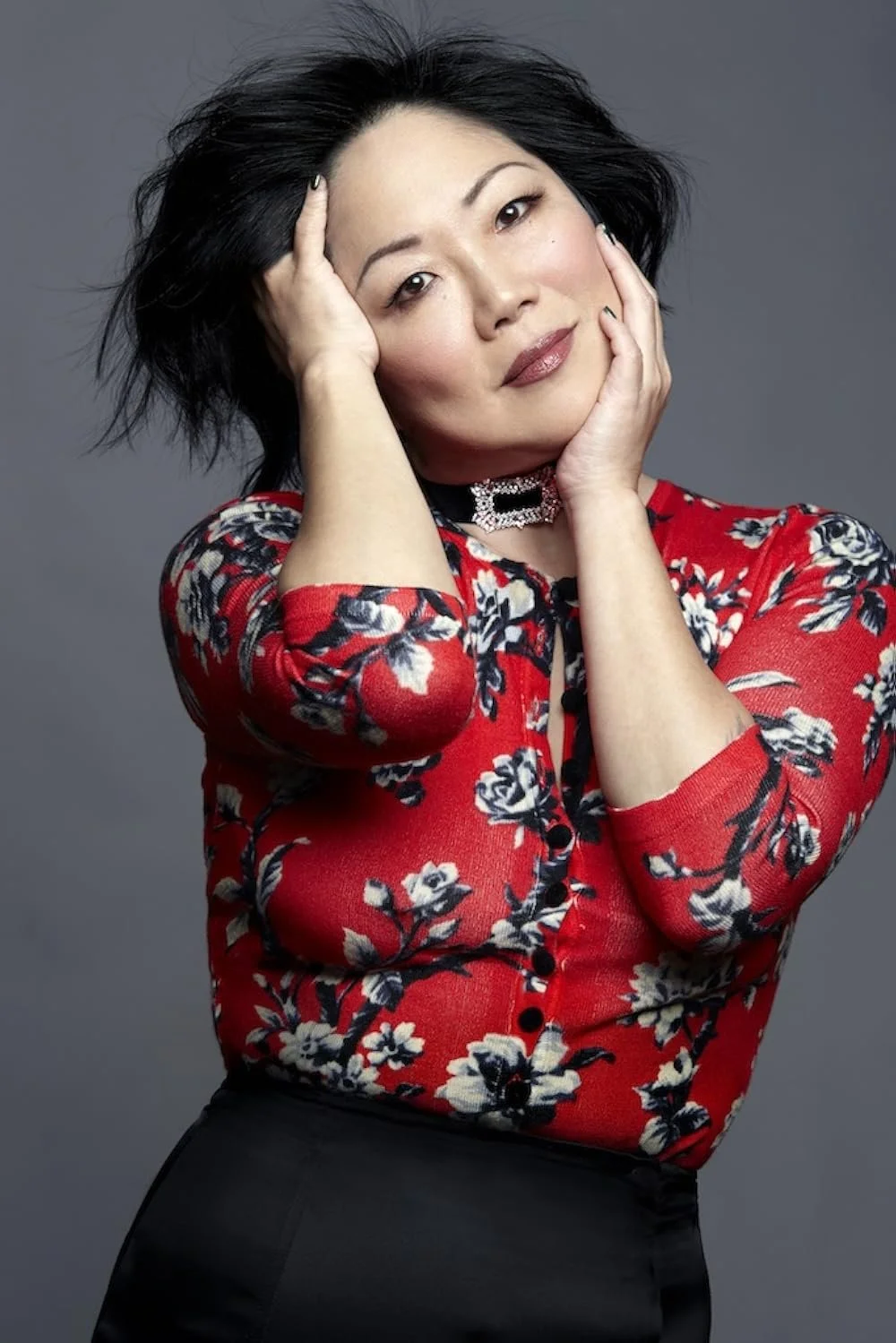 A woman with black hair in a floral red blouse posing with both hands touching her face against a gray background.