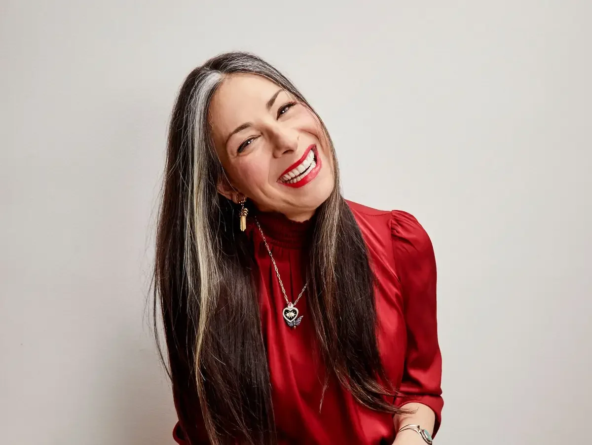 A woman with long dark hair and gray streaks, wearing a red satin blouse, red lipstick, hoop earrings, and a necklace with a black and white pendant, smiling and leaning slightly to her right against a plain light-colored wall.