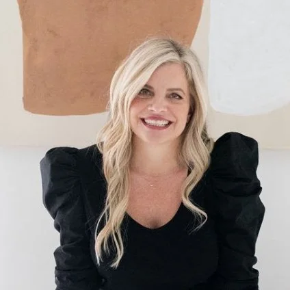 A smiling woman with long blonde hair wearing a black top with puffed sleeves, standing against an abstract multi-colored background.