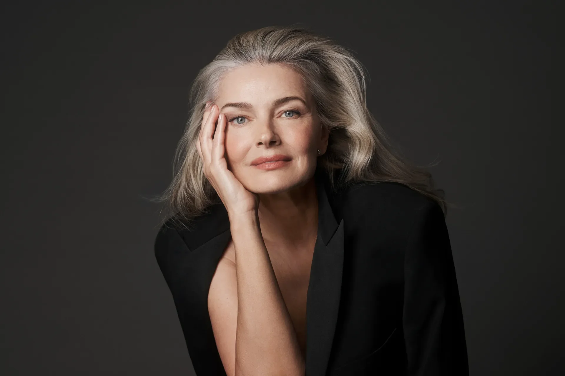A woman with silver hair and blue eyes, resting her face on her hand with a thoughtful expression, wearing a black blazer against a dark background.