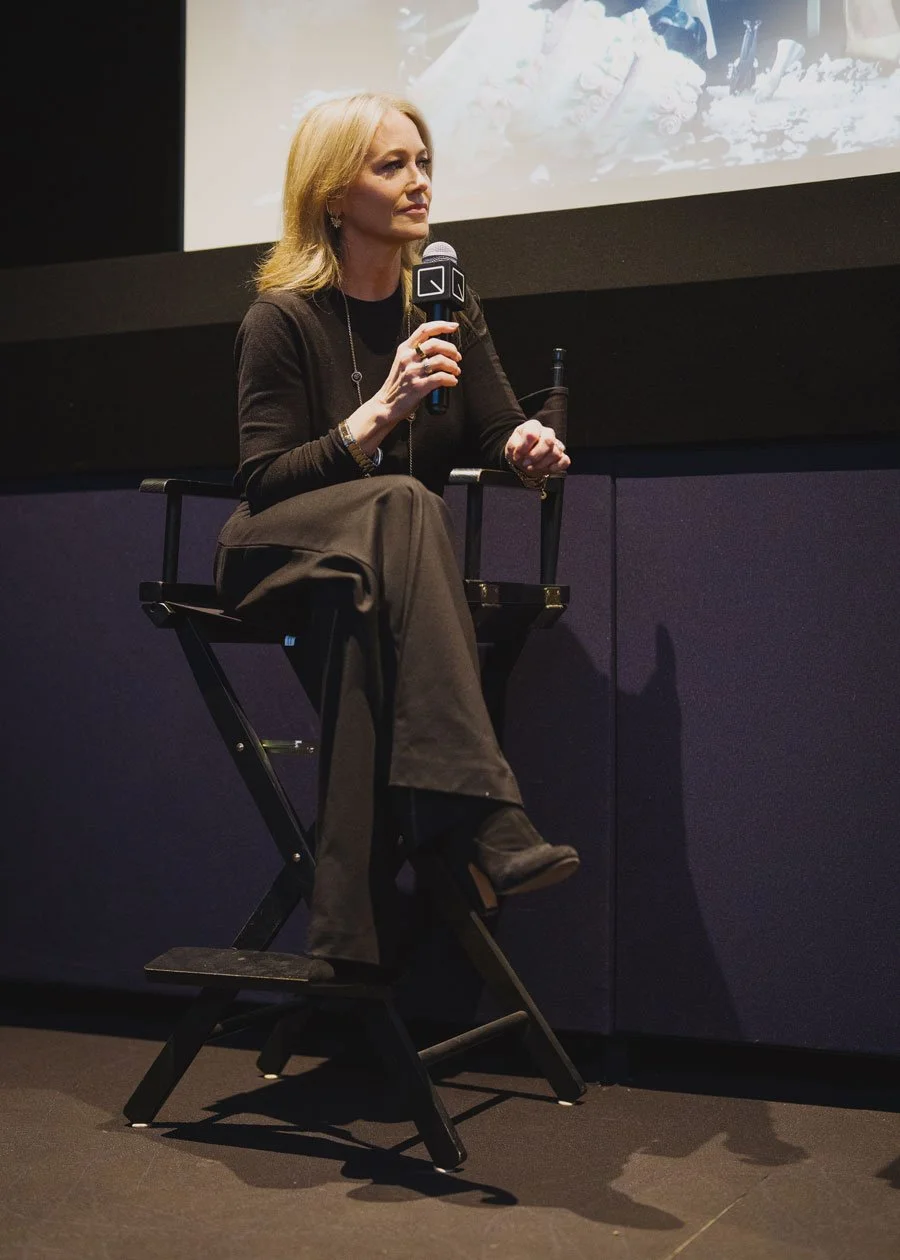 A woman with blonde hair sitting on a director's chair on a stage, holding a microphone, with a large screen behind her showing a presentation or conference.