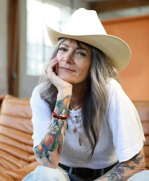 A woman with long hair and tattoos, wearing a white shirt and a cowboy hat, sitting indoors and resting her chin on her hand, smiling softly.
