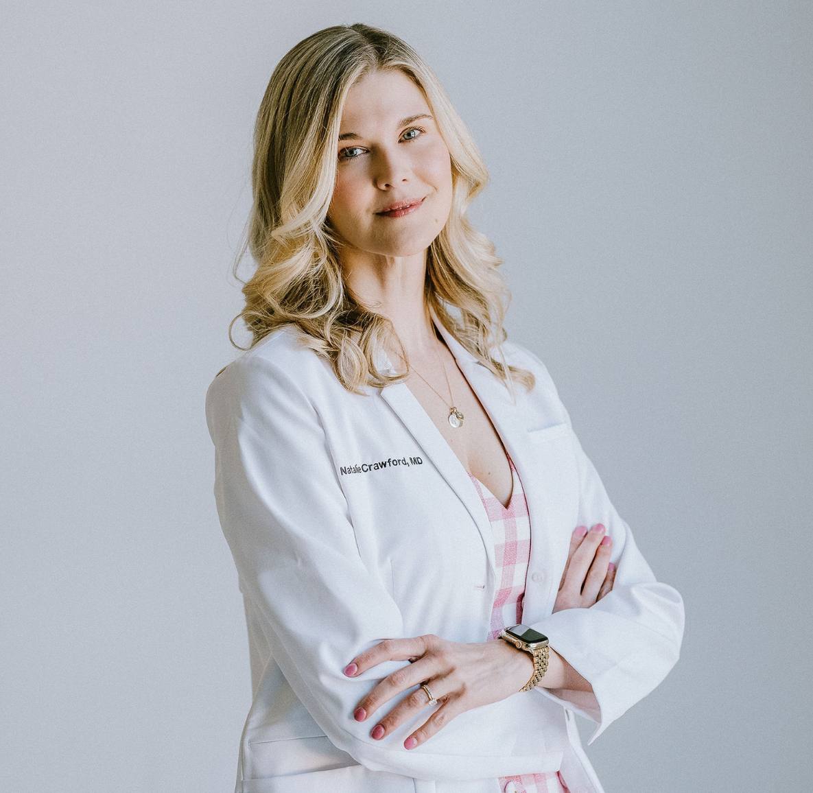 Professional woman with blonde hair wearing a white lab coat, crossed arms, and a pink checkered shirt, standing against a plain background.