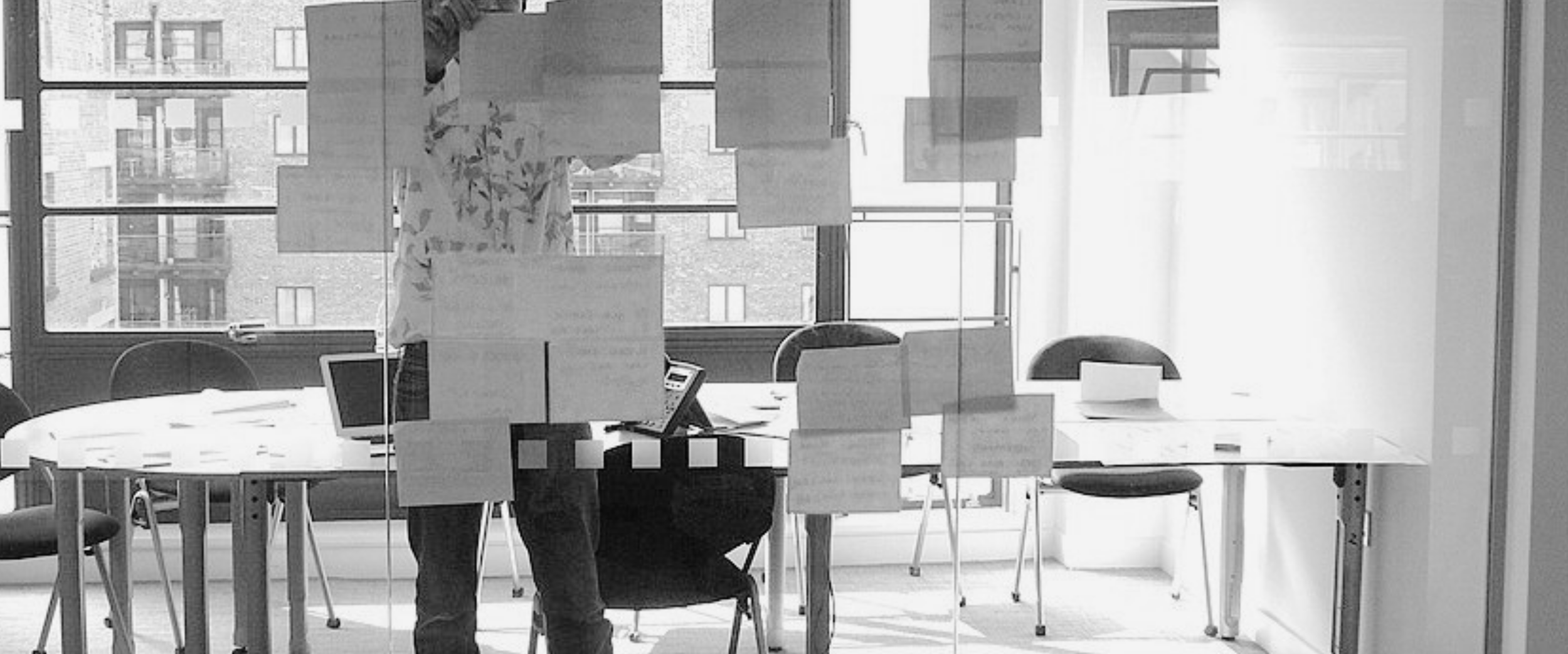 An empty office conference room with large windows, a round table, chairs, laptops, and papers on the table.