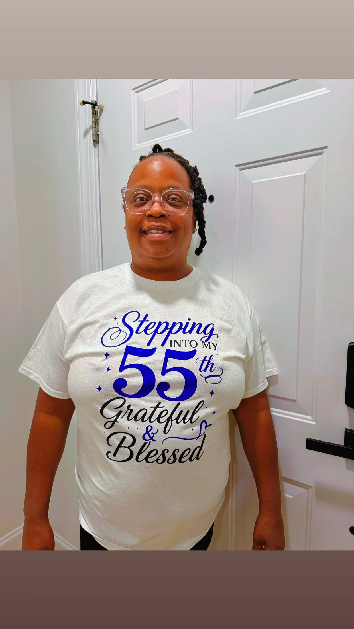 Woman smiling, wearing glasses and a white t-shirt with blue and black text celebrating her 55th birthday, standing in front of a white door.