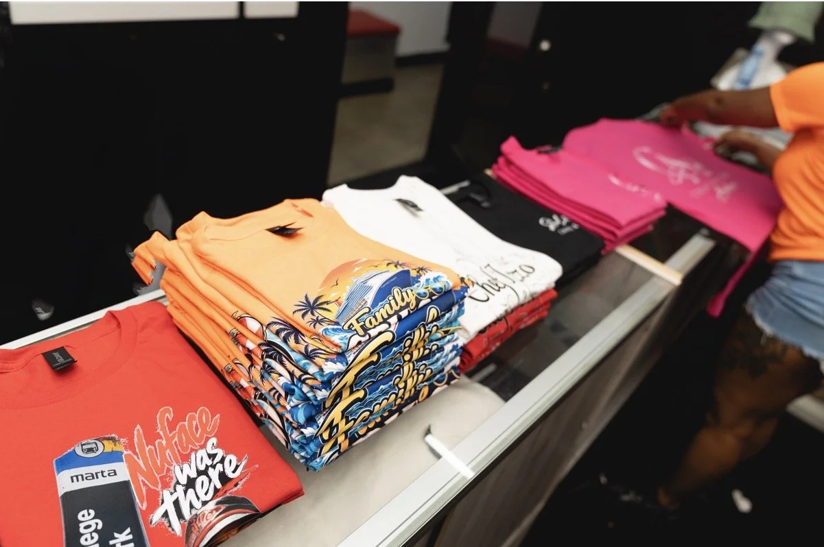 Stacks of colorful T-shirts on a display table, including red, white, orange, and pink, with various graphic designs.