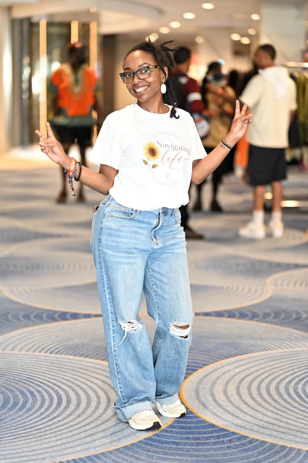 A young woman with glasses and dreadlocks smiling and making peace signs with both hands, standing in a busy indoor setting with people in the background.