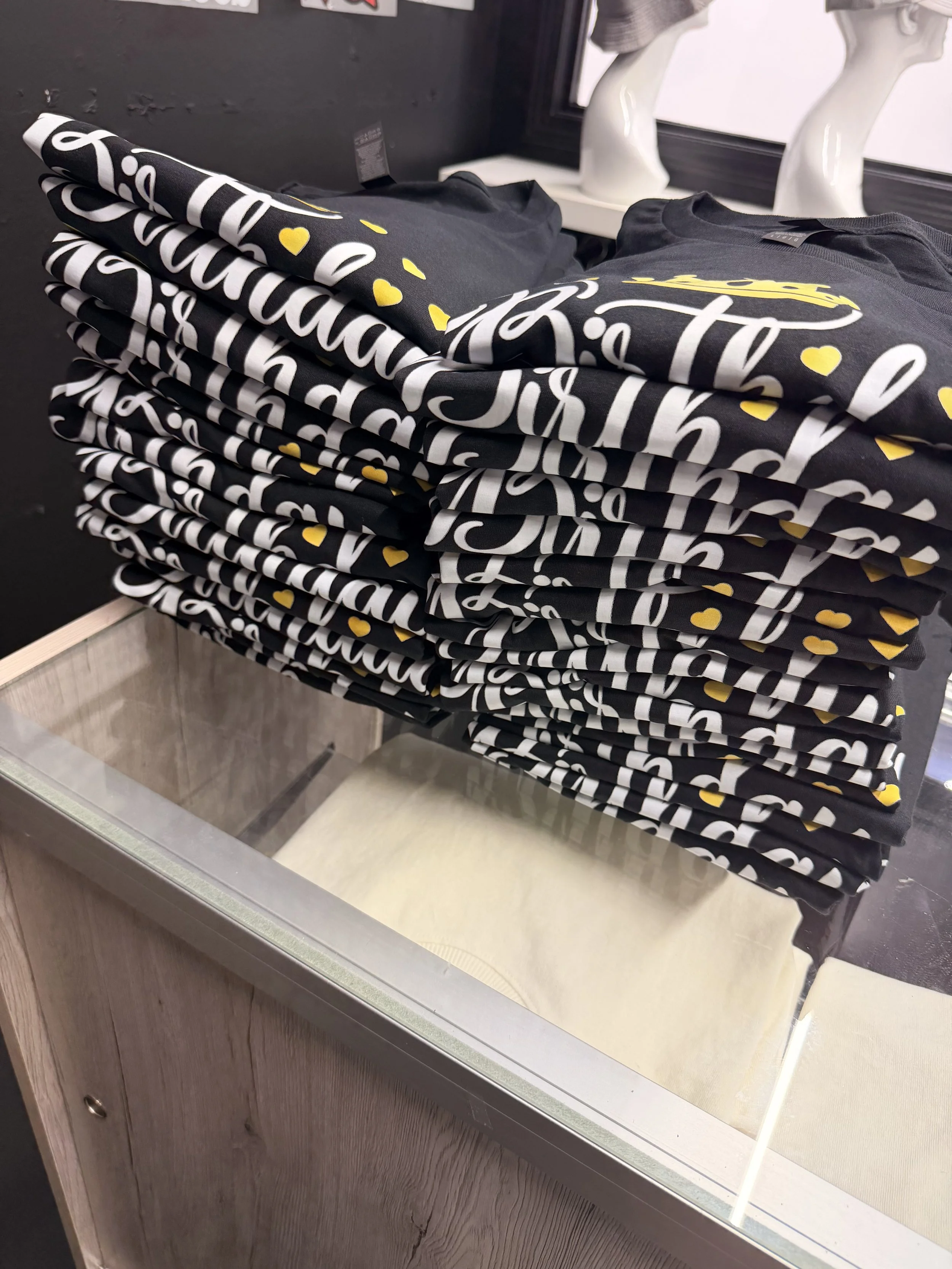 Stacked black T-shirts with white cursive text and yellow hearts on display at a retail store.