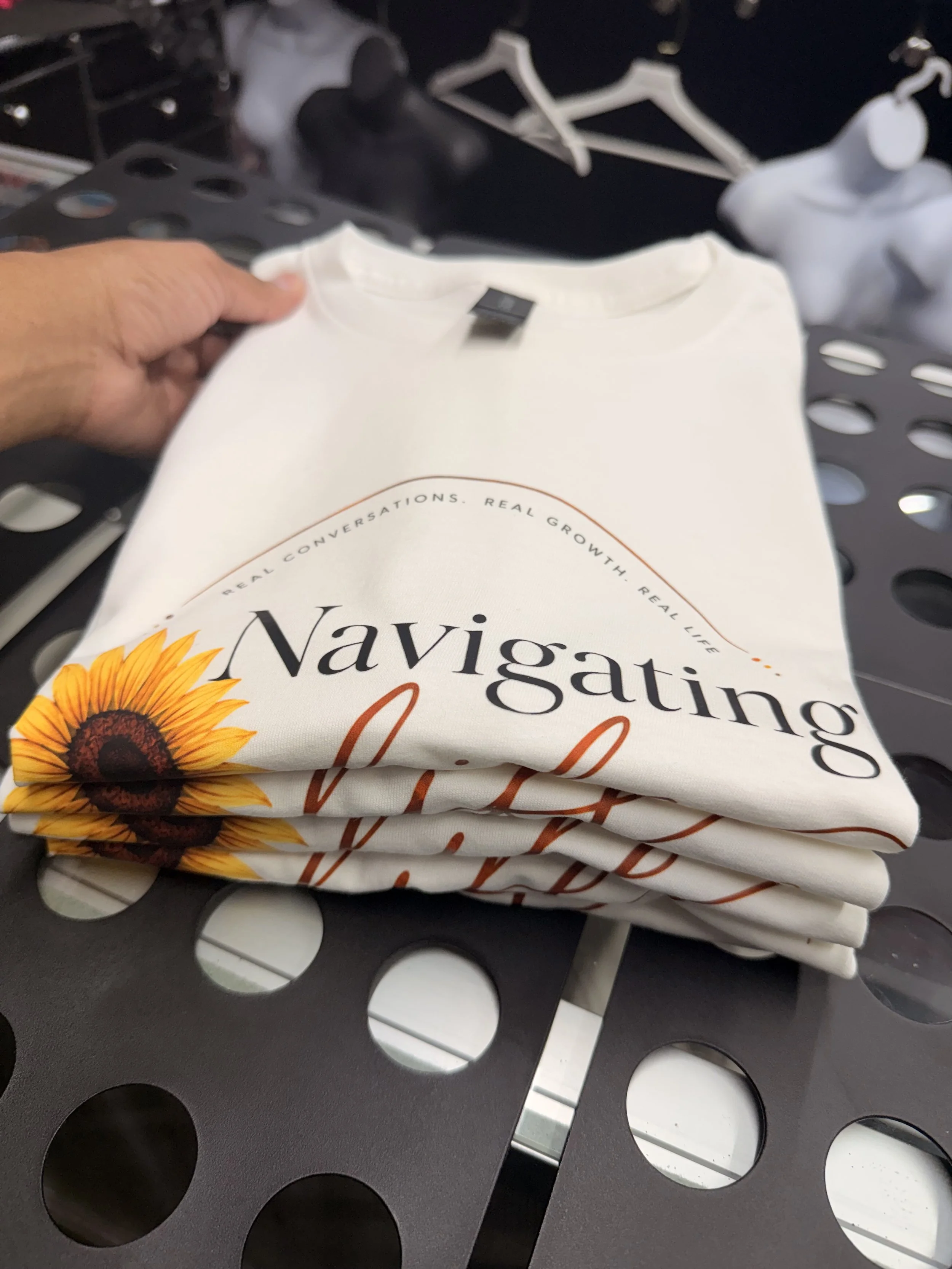 Stacked white T-shirts with sunflower graphic and text, placed on a black metal bench with circular cutout pattern.