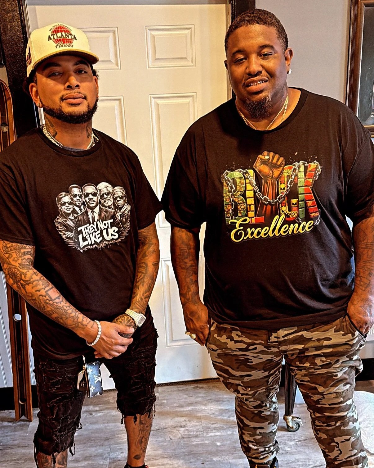 Two men standing indoors, both wearing black T-shirts with graphic designs and camouflage pants. The man on the left is wearing a baseball cap, has tattoos on his arms, and is holding his hands together. The man on the right has tattoos on his arms, he is wearing neck chains, and has his hand resting in his pocket.
