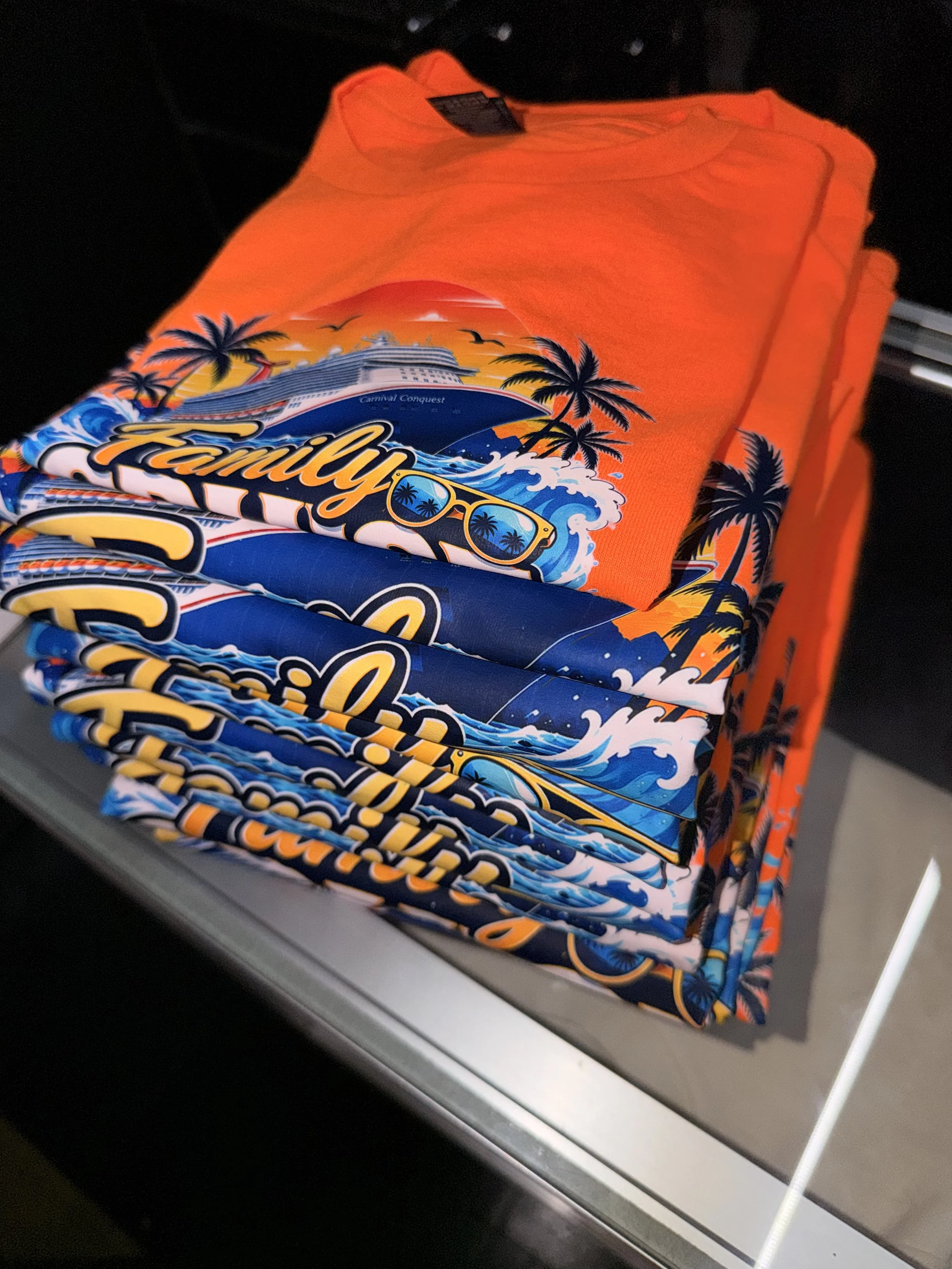 A stack of orange and blue towels with tropical and cruise ship designs, including palm trees, waves, and yachts, displayed on a store shelf.