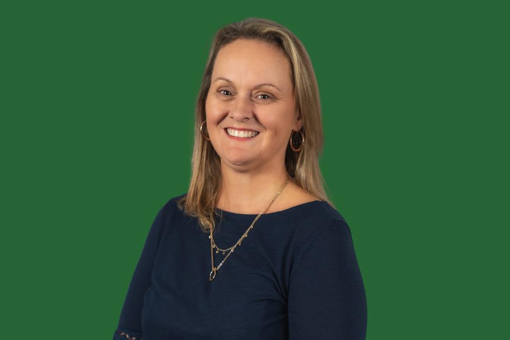 A smiling woman with blonde hair, wearing hoop earrings, a necklace, and a navy top, posing against a green background.