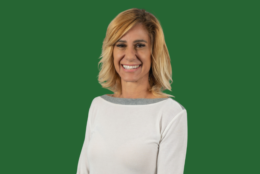 A woman with blonde hair smiling, wearing a white top with gray trim, standing against a green background.