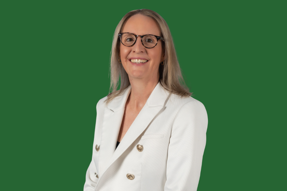 A woman with blonde hair, glasses, and earrings wearing a white blazer, smiling against a green background.