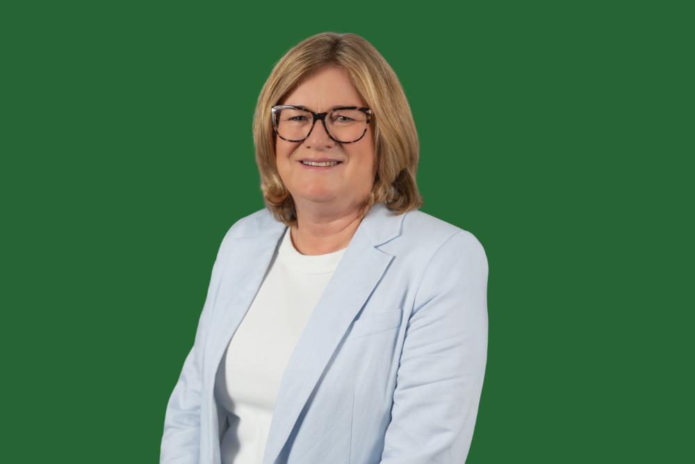 A smiling woman with blonde hair, wearing glasses, a light blue blazer, and a white shirt, smiling against a green background.