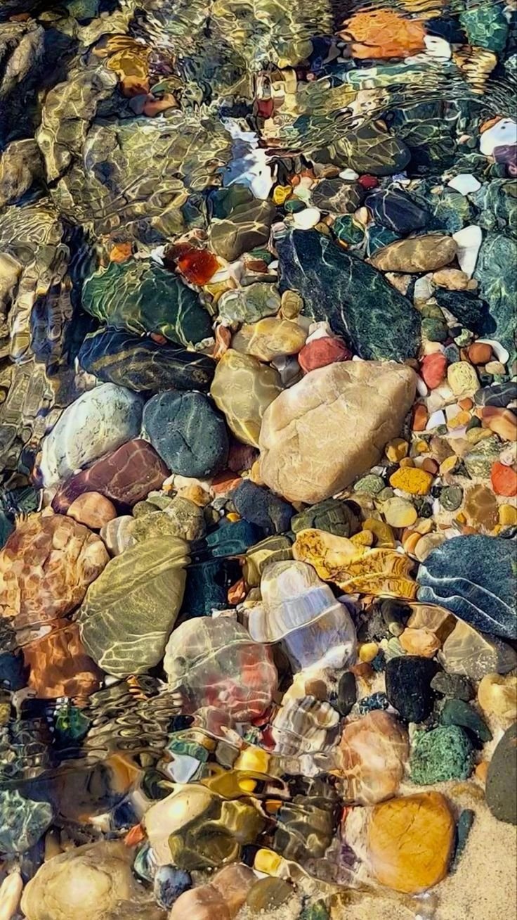 Colorful smooth stones and pebbles underwater in a shallow stream, with sunlight reflecting on the water's surface.