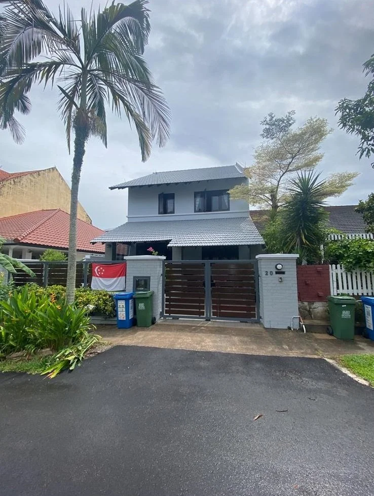 A two-story house with a gray roof, white walls, and a front gate, surrounded by greenery including palm trees, with a Singapore flag hanging on the front wall.
