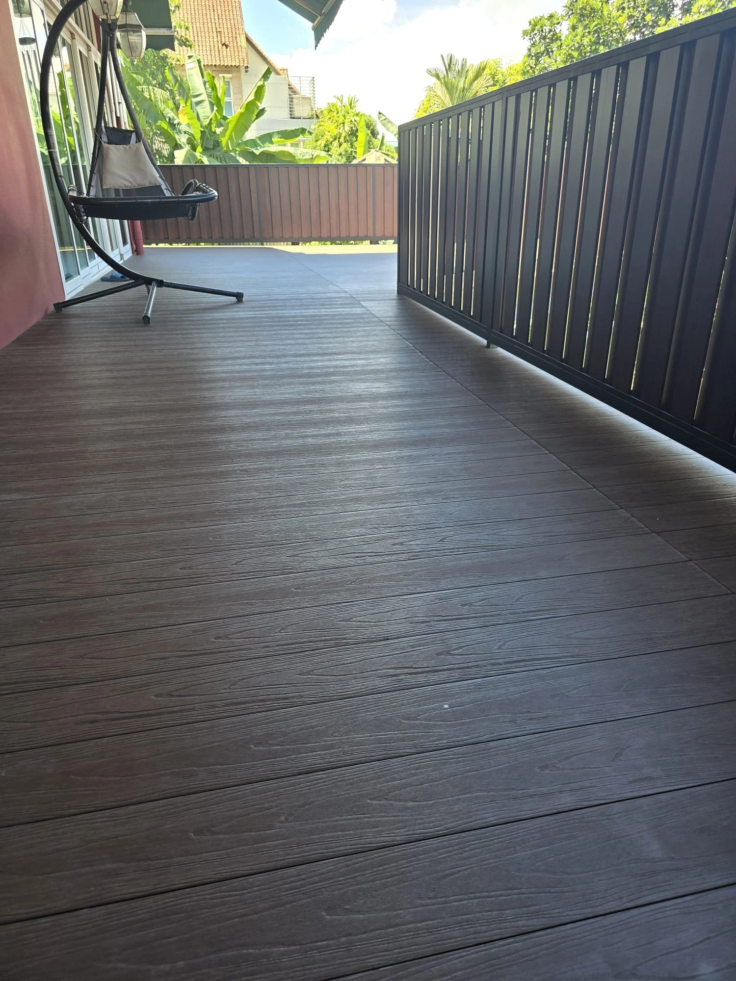 A balcony with wood grain flooring, a modern hanging chair, a large umbrella, and a view of green trees and neighboring houses in a bright, sunny day.