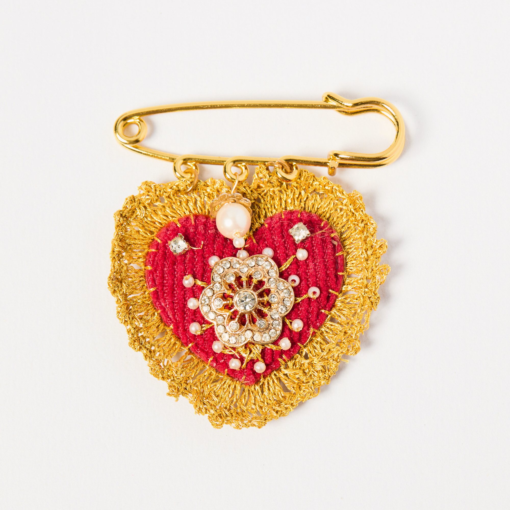 Gold safety pin with a decorative charm featuring red velvet, pearls, and rhinestones, attached to a gold chain against a white background.