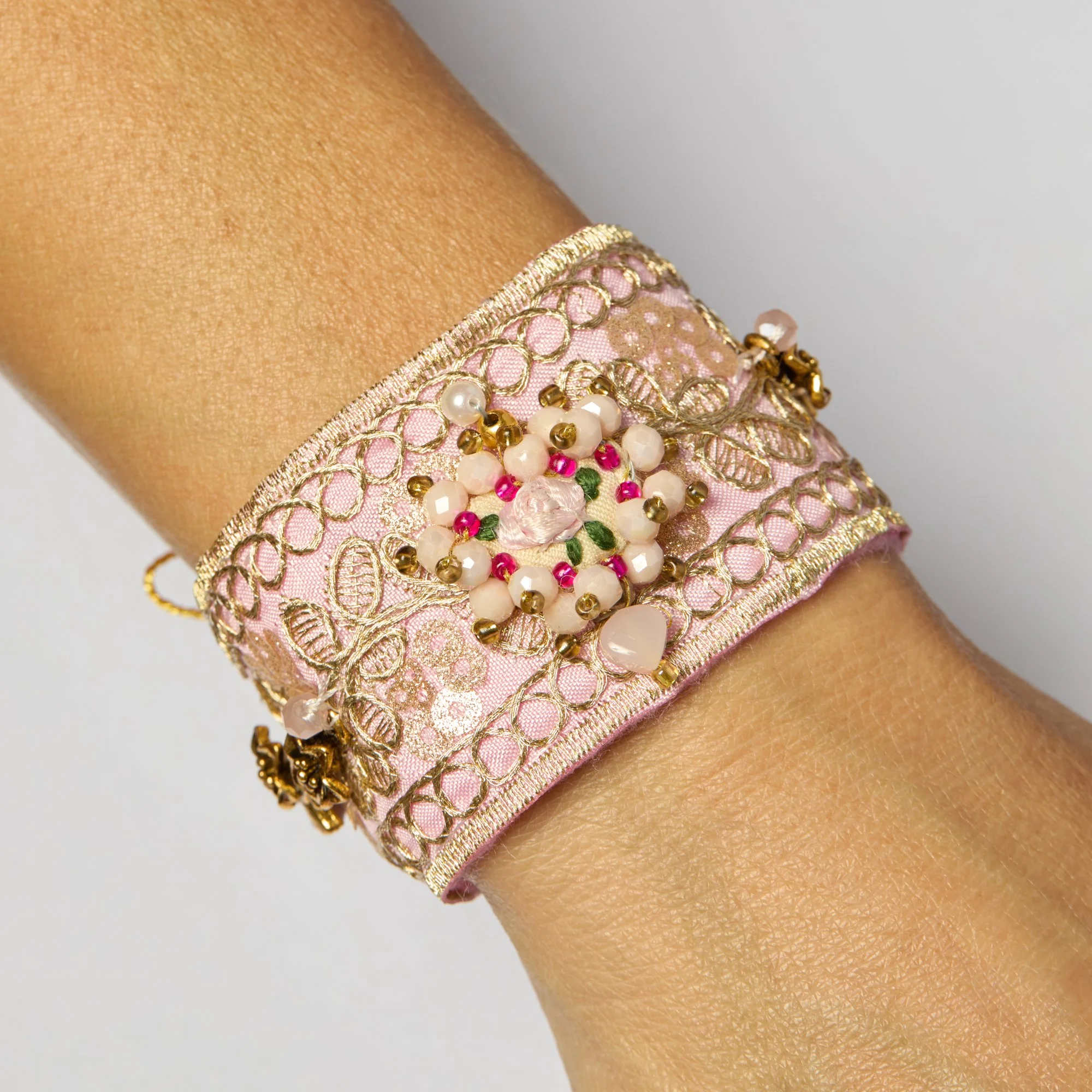 Close-up of a wrist wearing a pink fabric bracelet with extensive gold embroidery and beaded embellishments including pearls and pink beads.