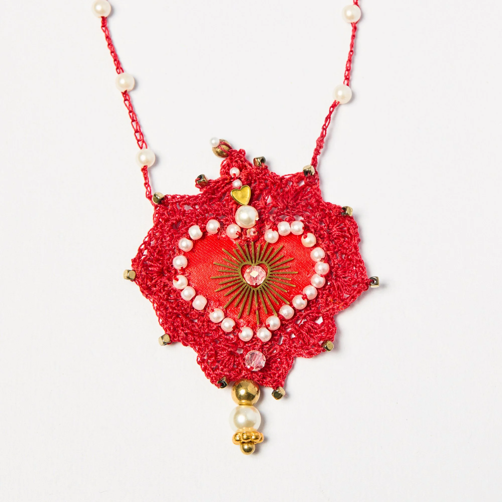 A red crocheted jewelry pendant with white pearl beads, gold accents, and a central gold-heart charm with a pink gemstone heart, hanging from a red thread with pearl beads.