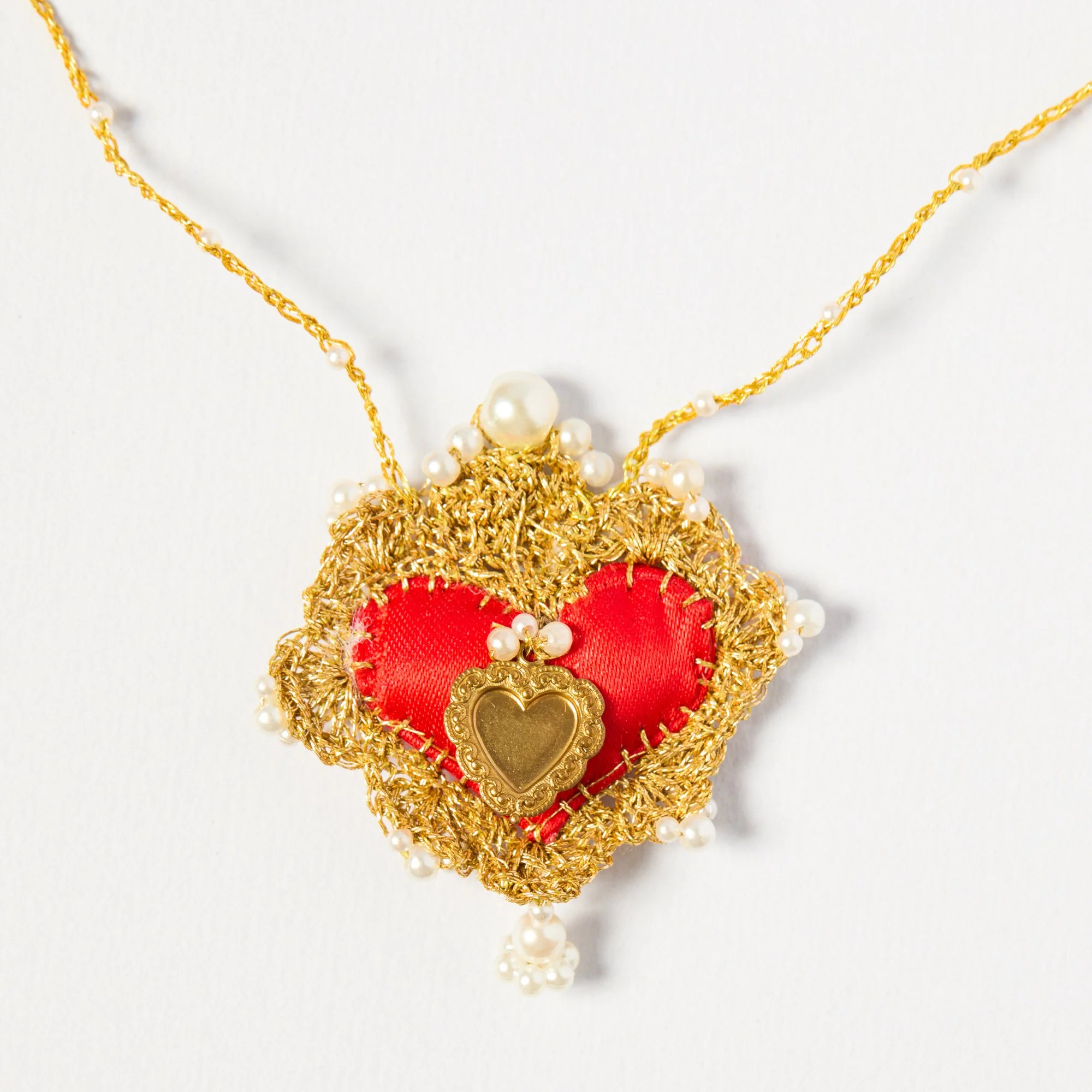 Close-up of a gold and pearl necklace with a decorative red fabric heart pendant and a gold heart charm, hanging against a white background.