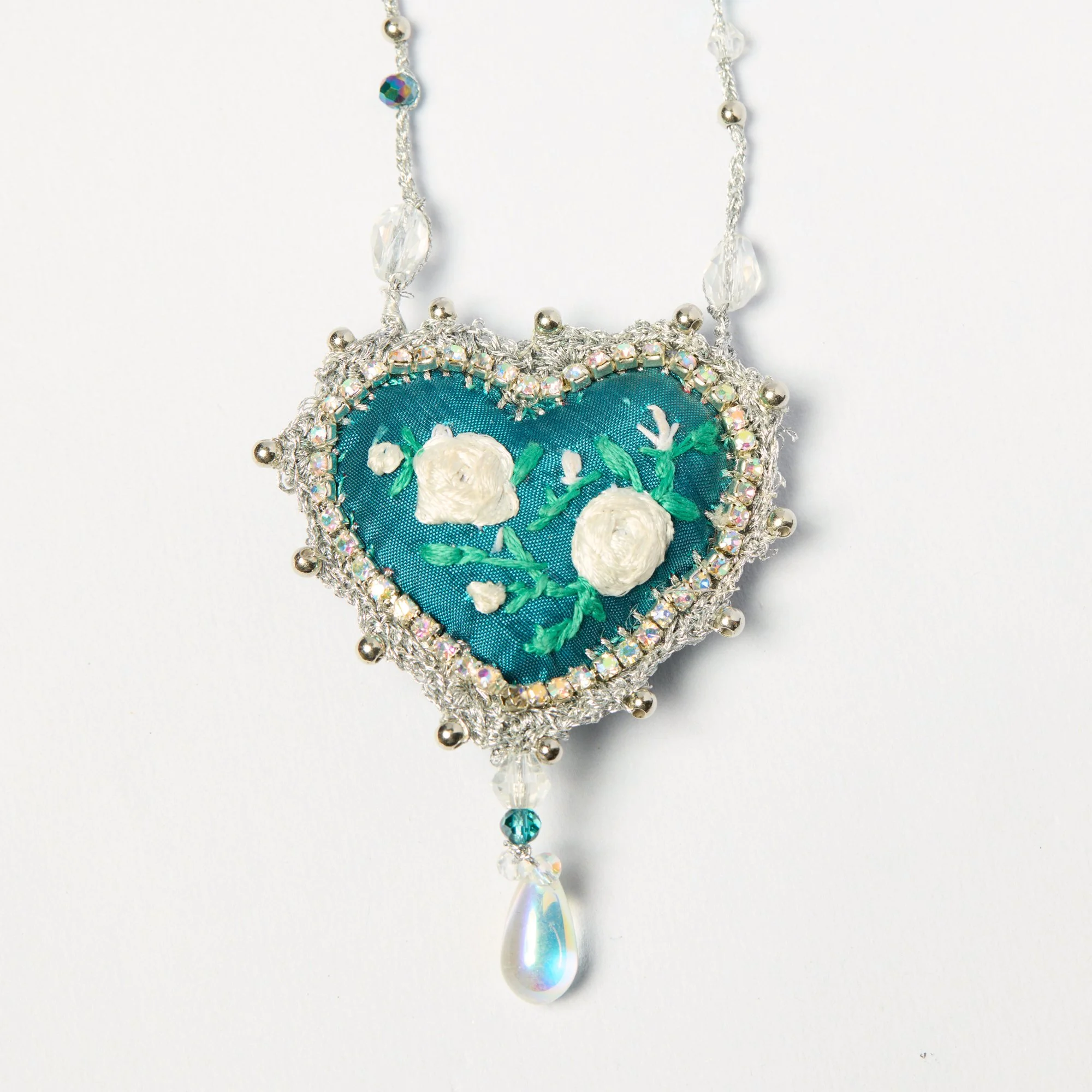 Close-up of a decorative necklace featuring a fabric heart with floral embroidery, surrounded by silver beads, rhinestones, and a dangling teardrop-shaped pearl.