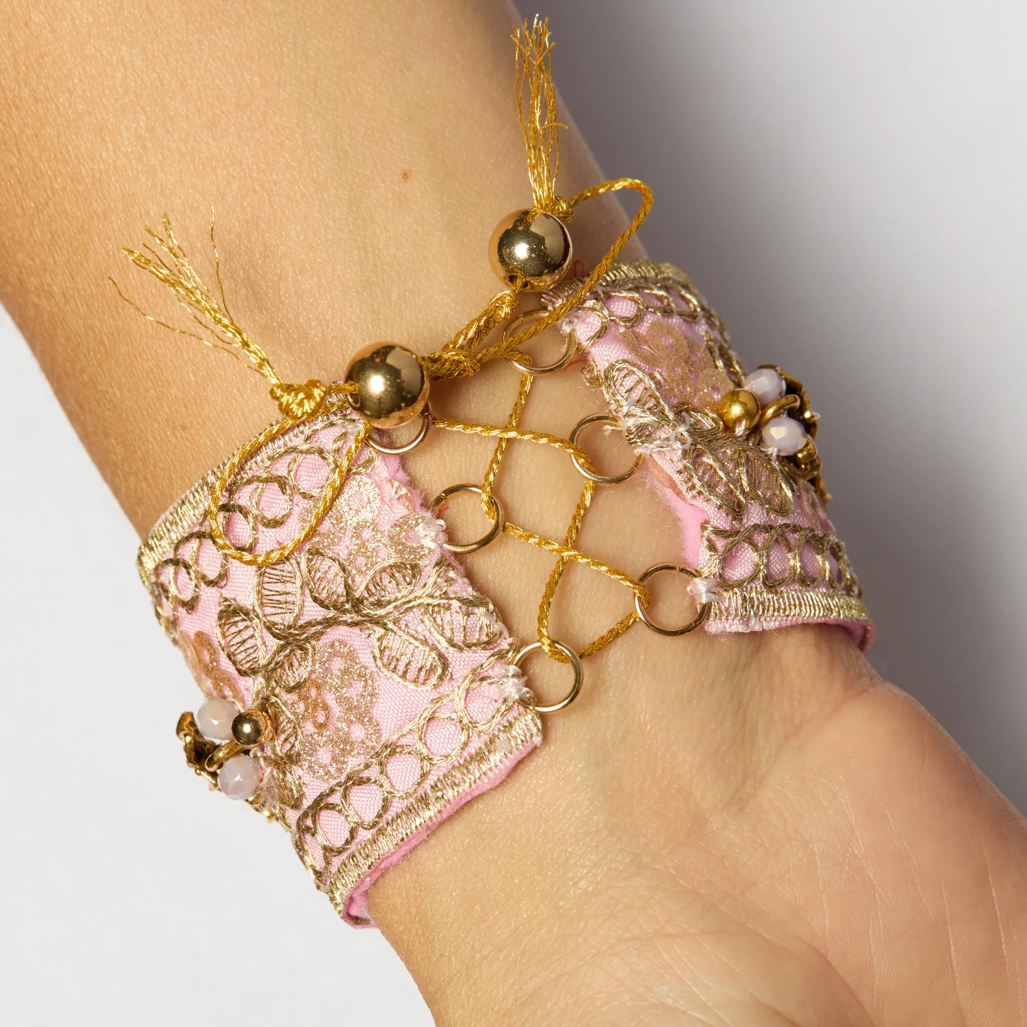Close-up of a person's arm adorned with multiple gold and pink bracelets featuring beads, chain links, and intricate lace-like fabric detailing.